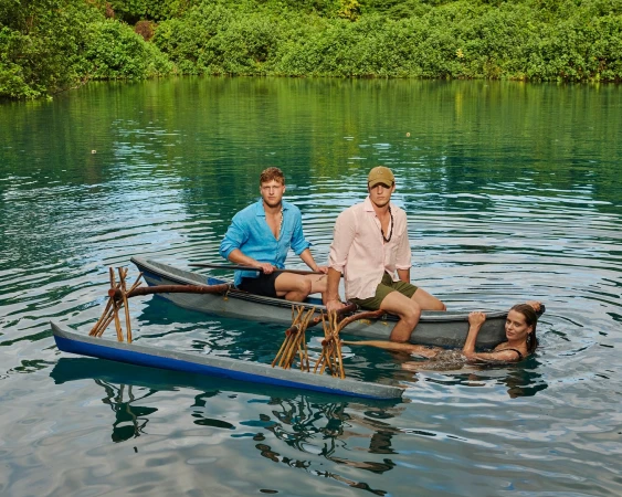 Three people in a canoe on a lake surrounded by lush greenery. Two adult men are in the canoe and one adult woman is in the water.