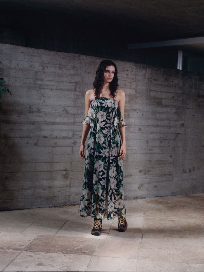 A young adult woman with long, dark hair poses in a floral-patterned dress and brown boots against a textured concrete wall in an indoor setting.