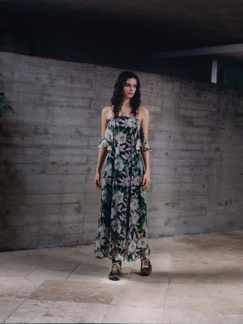 A young adult woman with long, dark hair poses in a floral-patterned dress and brown boots against a textured concrete wall in an indoor setting.