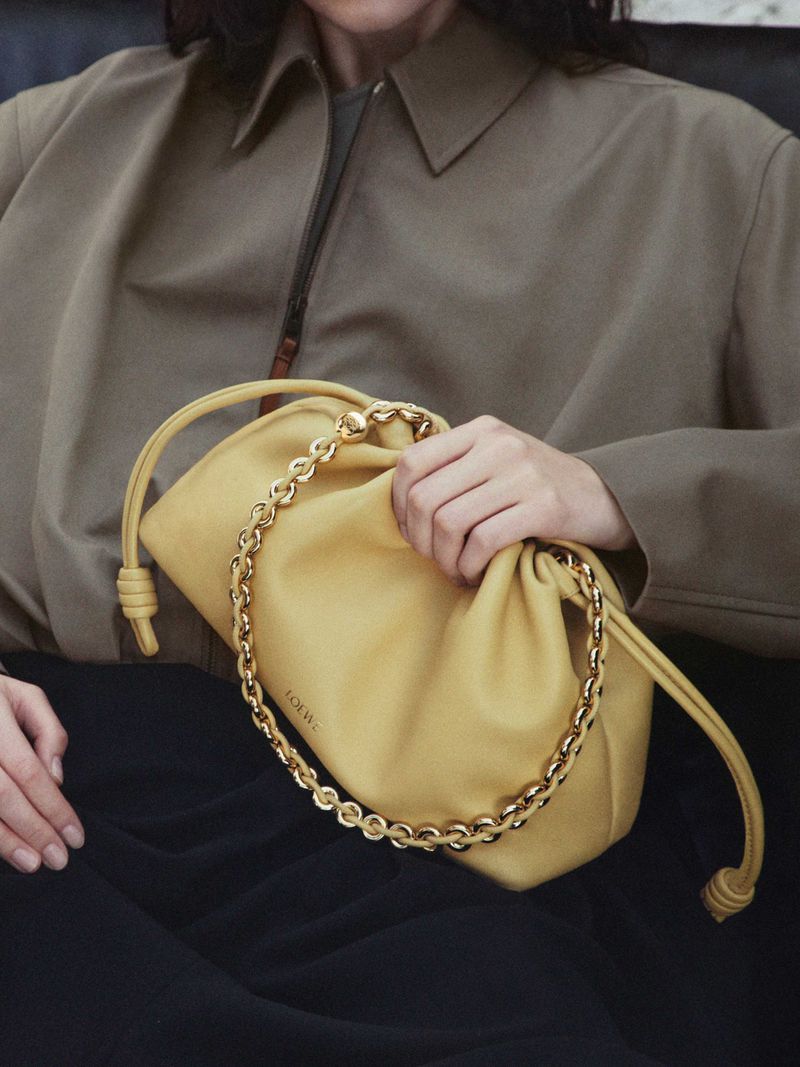 A pastel yellow Loewe Pouch bag with chain detail is held by an adult female in a medium shot, set against a neutral background.