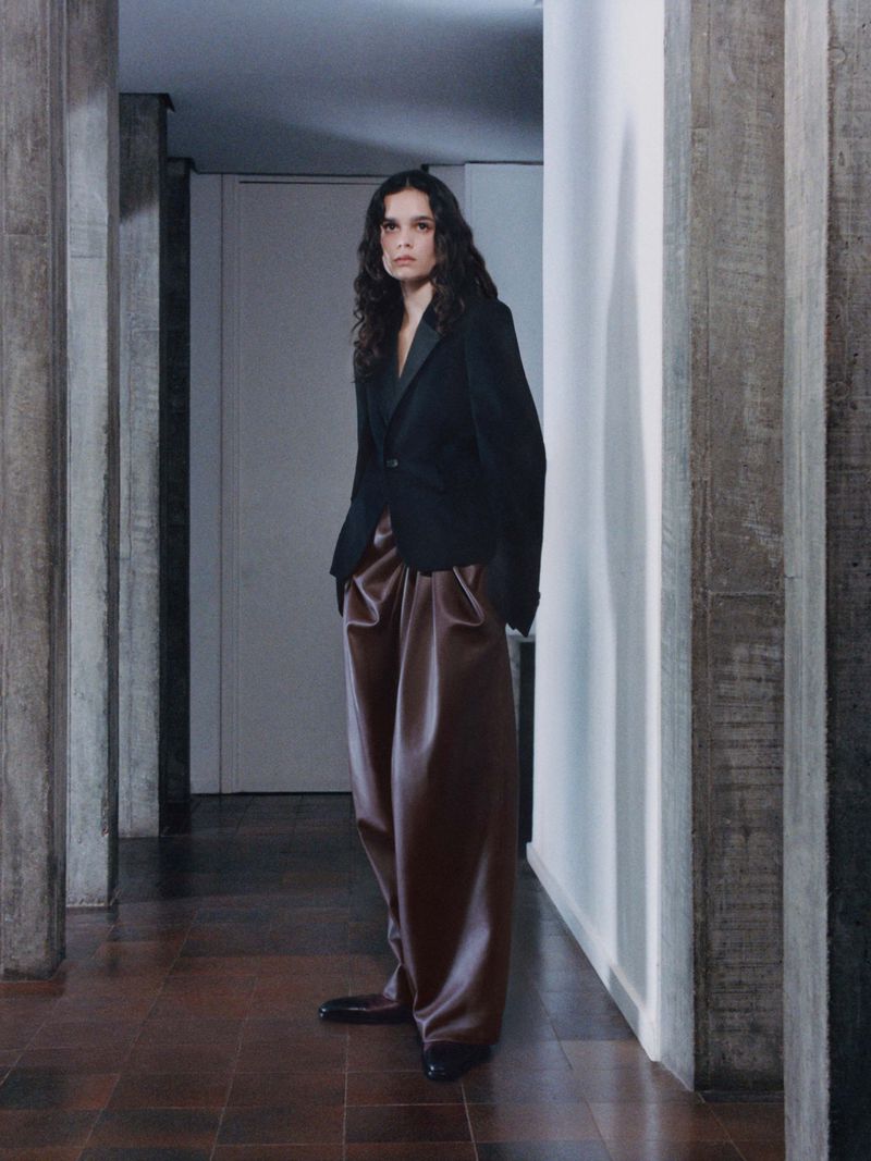 Fashion shot of a woman in a black blazer and wide-leg brown pants standing in an indoor hallway with a concrete pillar background.