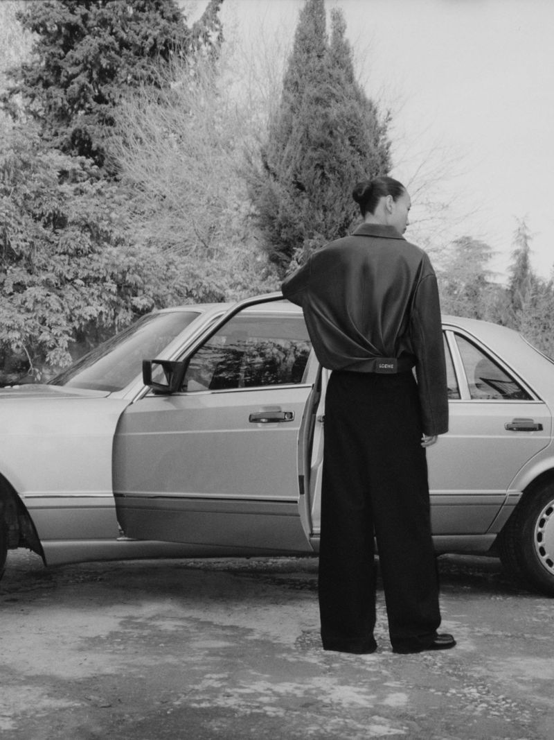 Monochrome shot of a female in a brown shirt and black pants standing next to a silver car in an outdoor setting with trees in background.
