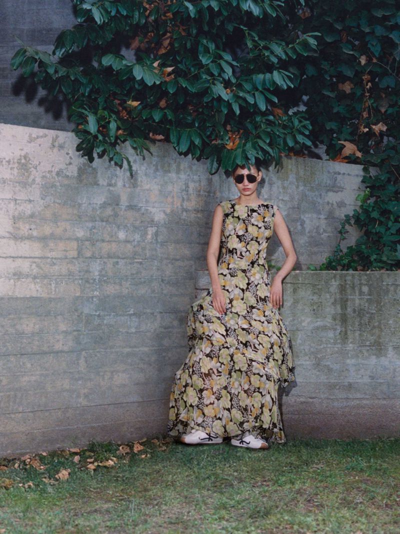 Model wearing a floral print maxi dress and sunglasses leaning against a weathered stone wall beneath green foliage.