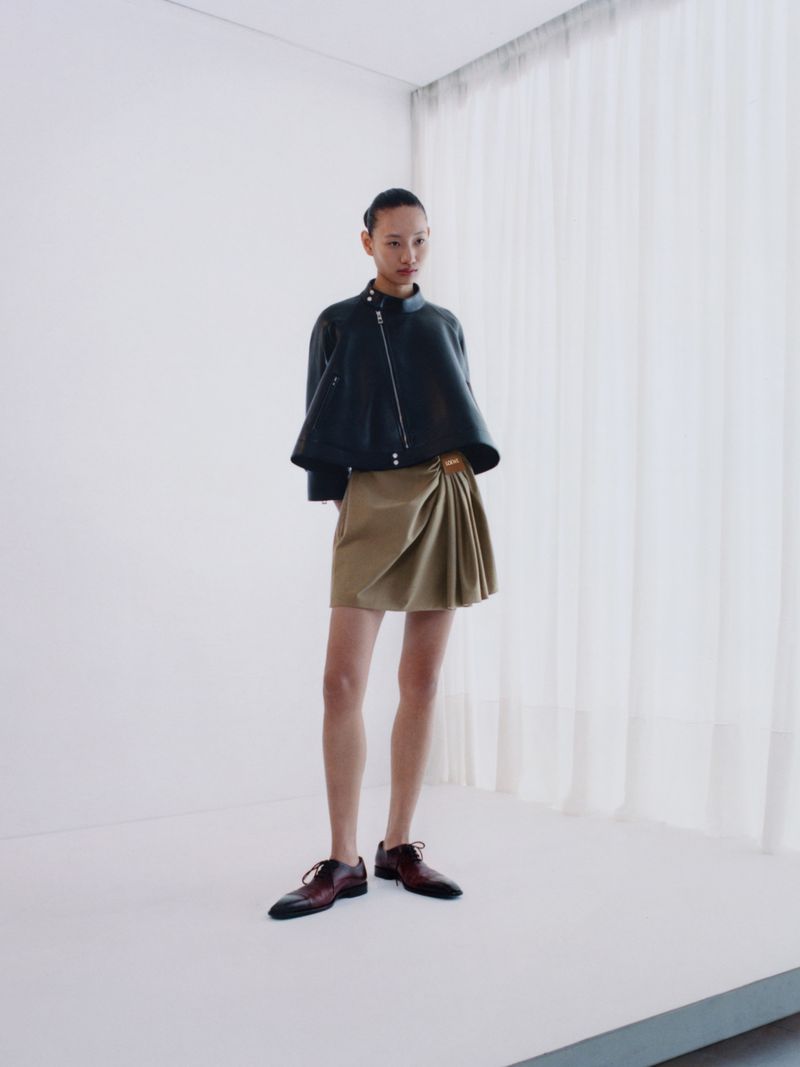 Fashion shot of an Asian adult woman in a dark jacket and tan skirt, standing in a white studio with soft lighting and subtle shadows.
