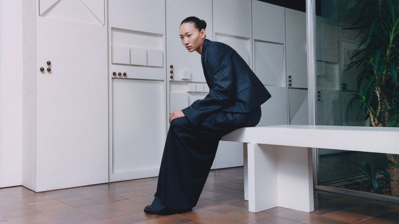 An Asian female model in a pinstripe suit sits on a white bench in a minimalist interior with plants. Fashion editorial photography.