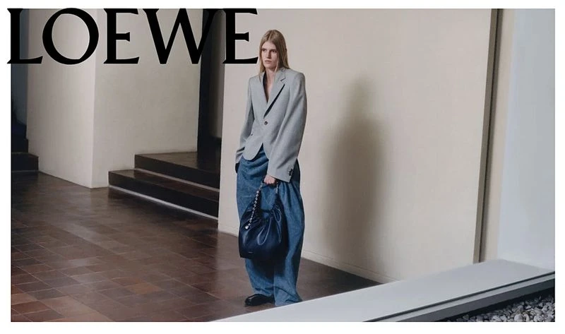 Fashion editorial shot of a woman in an oversized grey blazer and baggy jeans carrying a black bucket bag, posing in a minimalist interior.