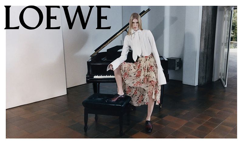 Fashion shot of a woman with long blonde hair posing in front of a piano, wearing a white top and floral print skirt, with Loewe logo.