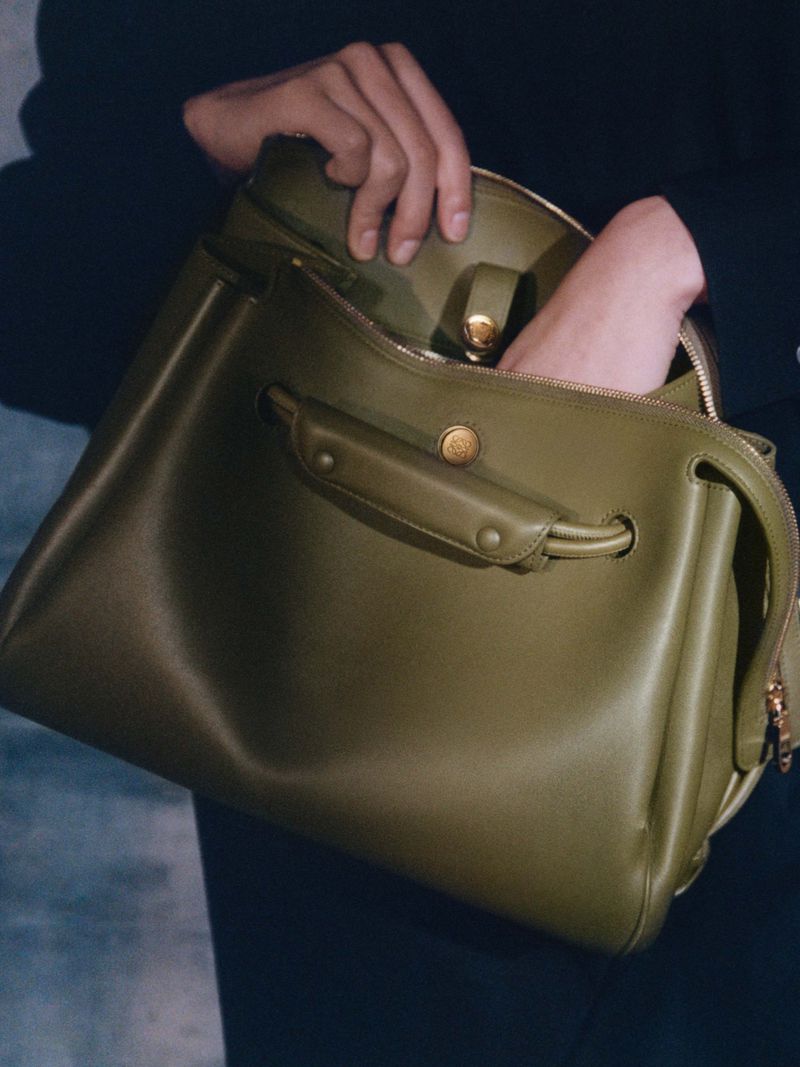 Olive green leather tote bag with gold hardware shown with hand opening the bag in a close up shot on a dark background.