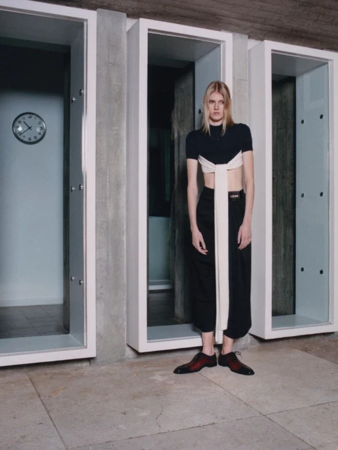 A slender woman in a two-tone outfit stands in front of storage units, shot in eye-level medium shot. A clock is visible on the wall.