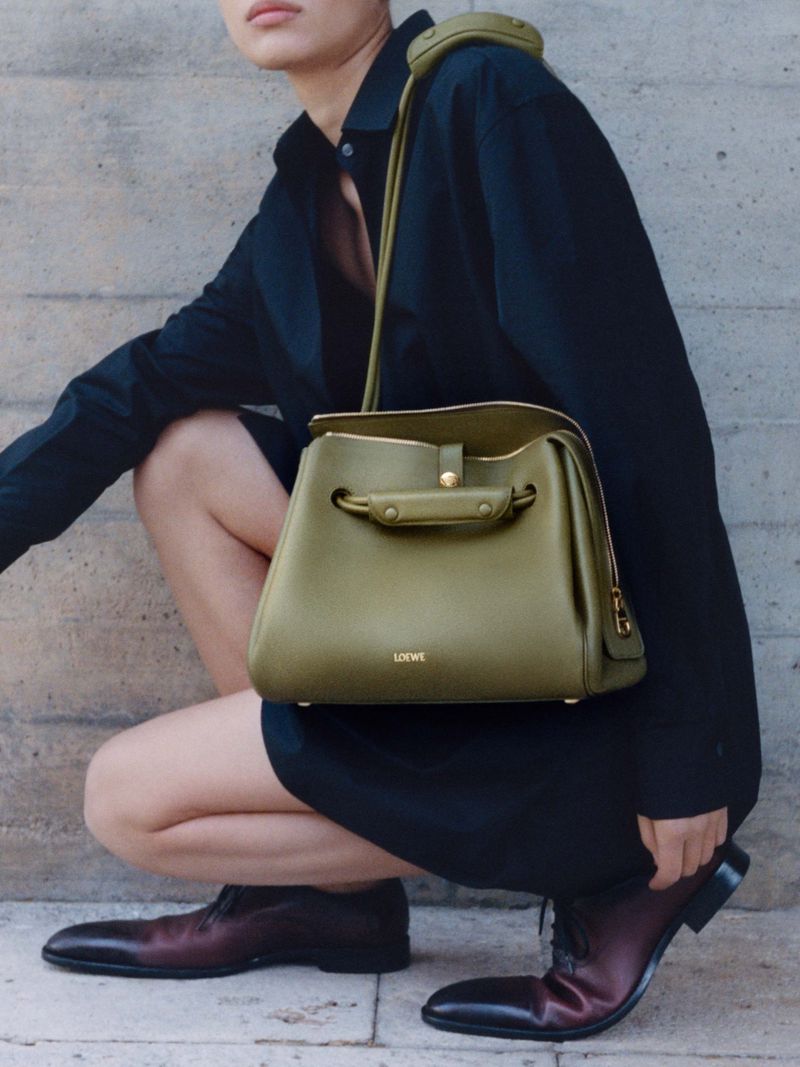 Olive-green leather satchel bag with top handles and shoulder strap, worn by an adult female wearing a dark dress and brown shoes.