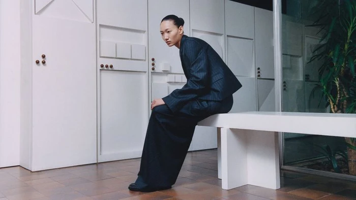 Model wearing dark suit seated on a white bench against a minimalist interior background for Loewe