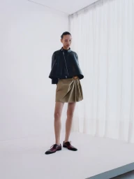 Model standing on a white surface wearing a dark leather jacket and beige skirt against a white curtain background.
