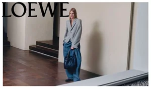 Model wearing a grey blazer and blue trousers holding a dark handbag inside a building with tiled floors and stairs.