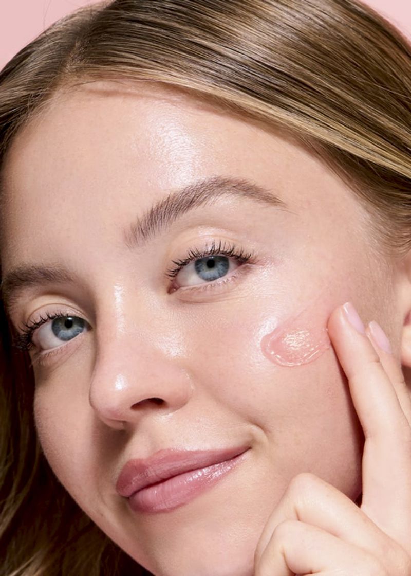 Close-up studio shot of a young woman with blonde hair smiling as she touches her cheek displaying a pink skincare product.
