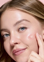 Close-up portrait of Sydney Sweeney applying pink skincare product to her cheek against a pink background
