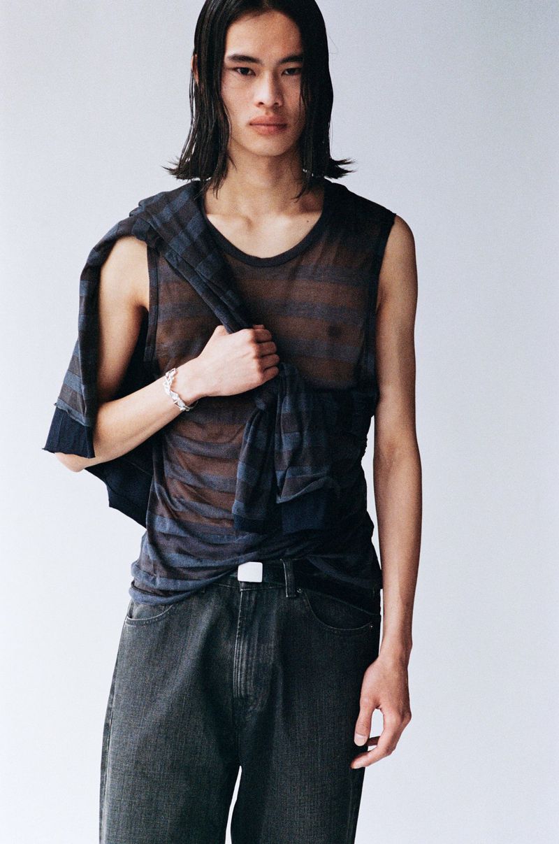 A slender, dark-haired Asian man poses in a sheer dark top and jeans, with a patterned jacket draped over his shoulder against a white backdrop.