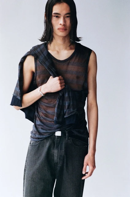A slender, dark-haired Asian man poses in a sheer dark top and jeans, with a patterned jacket draped over his shoulder against a white backdrop.