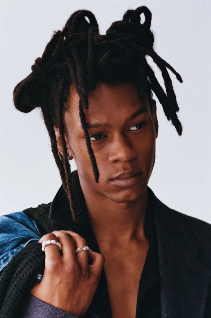 Portrait of a young Black man with dreadlocks wearing a black blazer, rings, and a denim jacket against a white background.