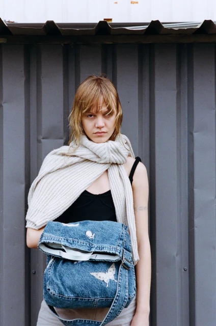 A woman stands outdoors against a metal wall, wearing a black top, grey bottoms, and denim bag with white butterfly and fish designs.