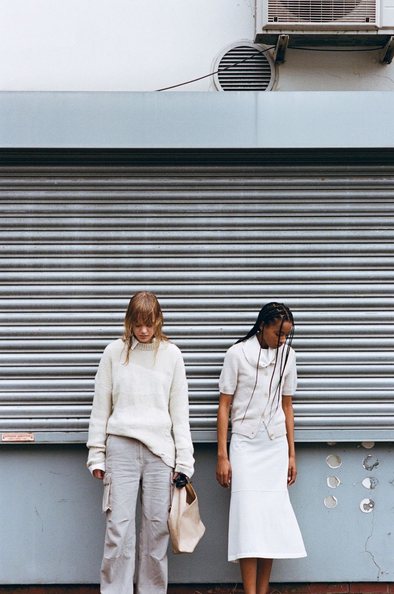 Two women in fashionable neutral-toned clothing pose against a metal industrial building exterior with an AC unit overhead.