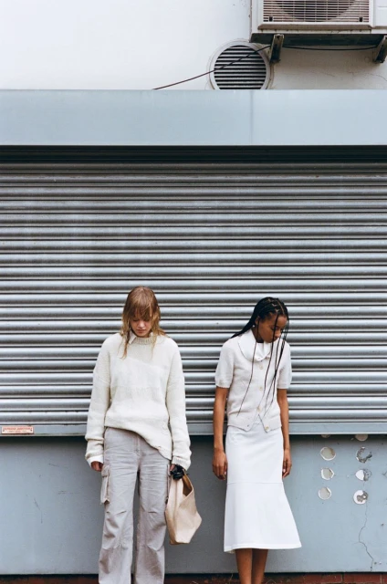 Two women in fashionable neutral-toned clothing pose against a metal industrial building exterior with an AC unit overhead.