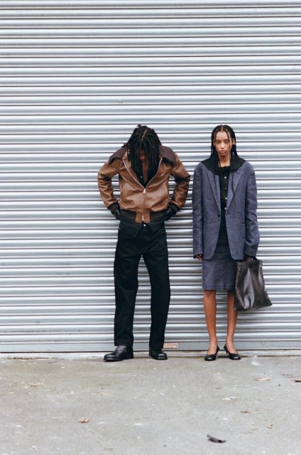 Two people wearing fashionable, formal attire pose against a corrugated metal wall with neutral expression; outdoor setting and natural light.