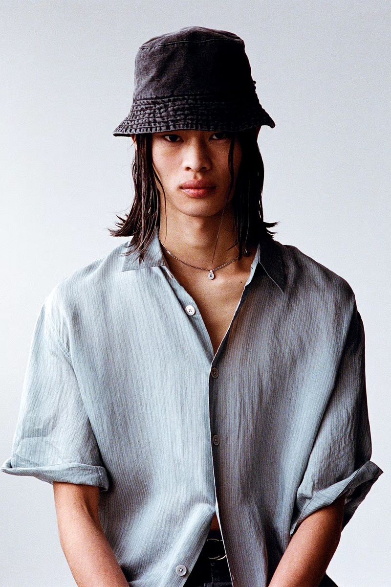 A young male model with long dark hair wearing a bucket hat and light blue shirt against a white backdrop.