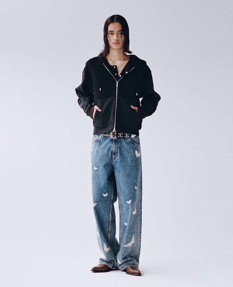 A slender man wearing a black zip-up jacket and butterfly-patterned jeans stands against a white backdrop in a full-length studio shot.
