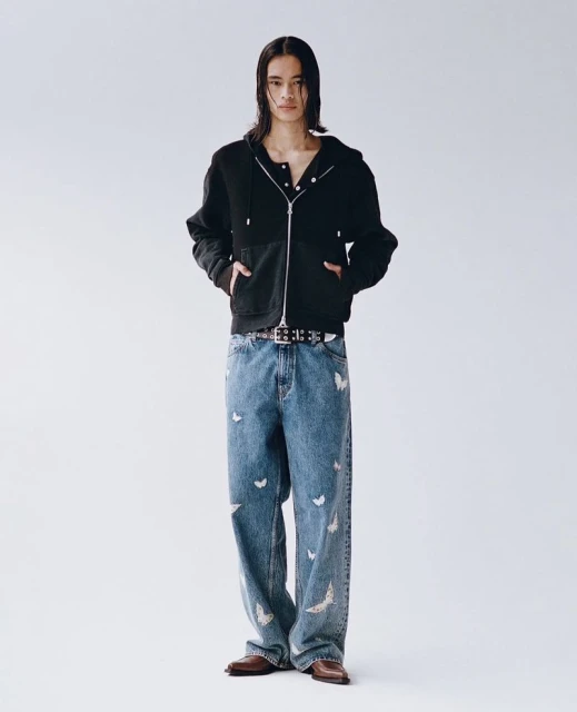 A slender man wearing a black zip-up jacket and butterfly-patterned jeans stands against a white backdrop in a full-length studio shot.