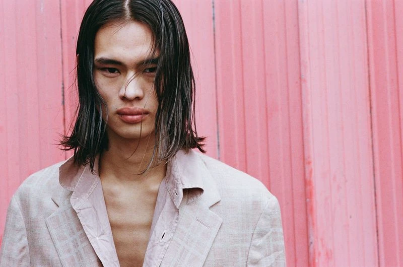 Close-up of a young adult male with long hair wearing a light blazer, posing against a pink corrugated metal wall.