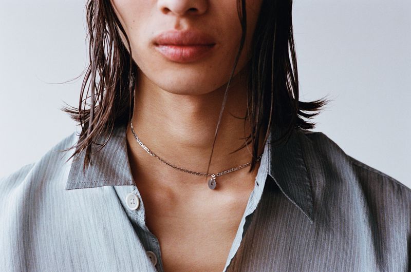 Close-up of a young adult wearing a light blue striped shirt and silver necklace, gazing straight ahead in a brightly lit, neutral setting.