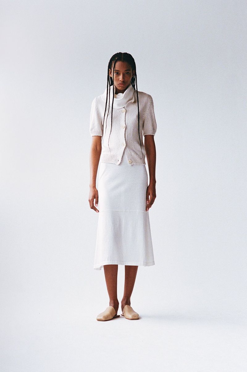 A woman stands in a photo studio wearing a light-colored sweater, white skirt, and tan flats, looking directly at the camera with a neutral expression.