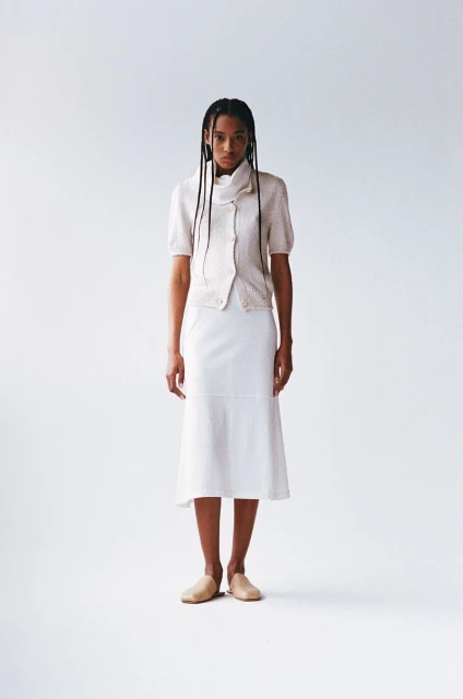 A woman stands in a photo studio wearing a light-colored sweater, white skirt, and tan flats, looking directly at the camera with a neutral expression.