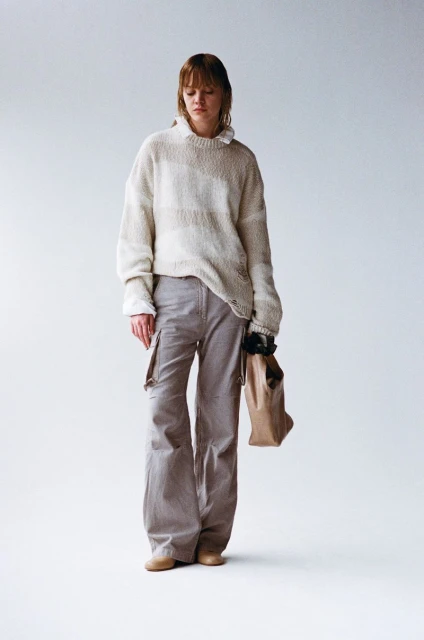 A full shot of a young woman with bangs in an oatmeal sweater and gray cargo pants, holding a brown paper bag in a photo studio.