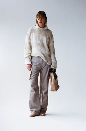 A full shot of a young woman with bangs in an oatmeal sweater and gray cargo pants, holding a brown paper bag in a photo studio.