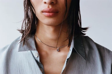 Close-up portrait of a model wearing a grey collared shirt and silver chain necklace against a white background.