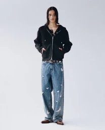 Model wearing a black zip hoodie and patterned blue jeans with hands in pockets against a plain grey background