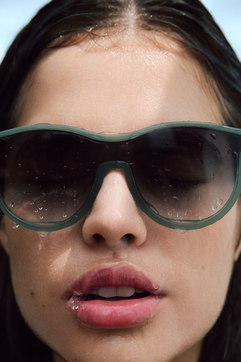 Close-up of a young woman's face, wearing sunglasses with water droplets, wet dark hair, and red lipstick in bright, diffused lighting.