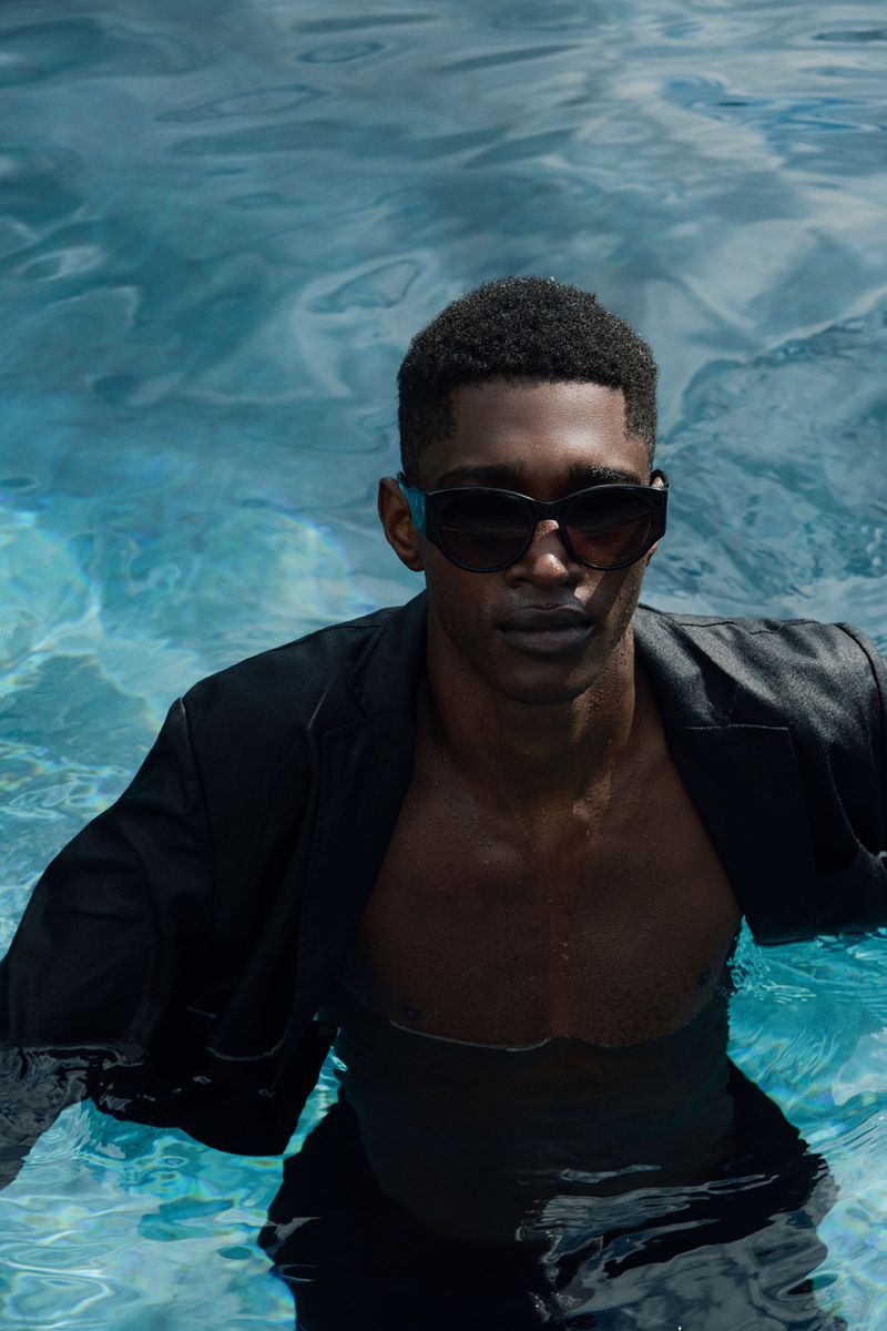 A Black male model stands waist-deep in a pool wearing sunglasses and a black suit jacket, looking at the camera with a serious expression.