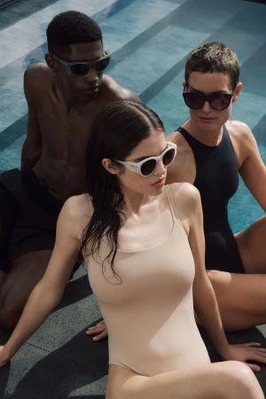 Three models in swimsuits and sunglasses pose near a pool, showcasing summer fashion and swimwear trends against a bright, outdoor setting.