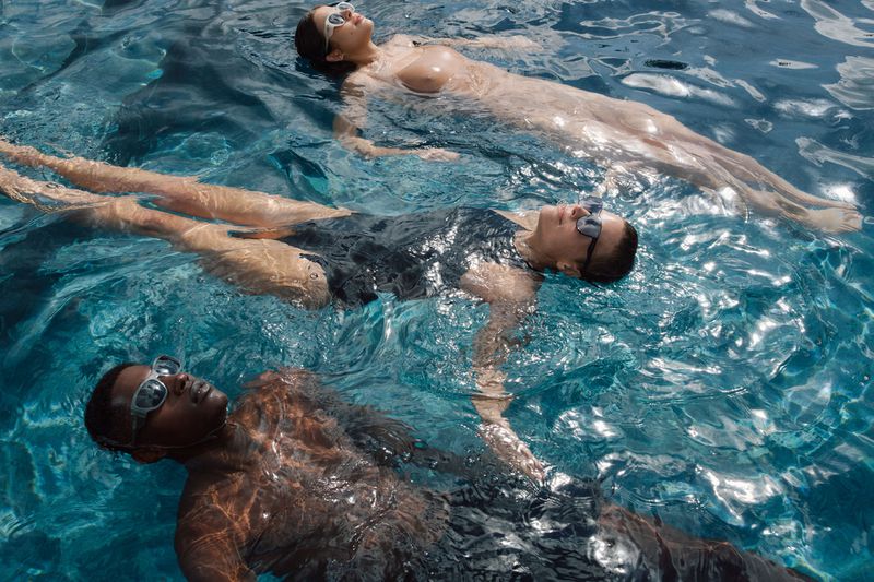 Three people float in blue water. One African-American man and two white women are submerged in a swimming pool on a sunny day.