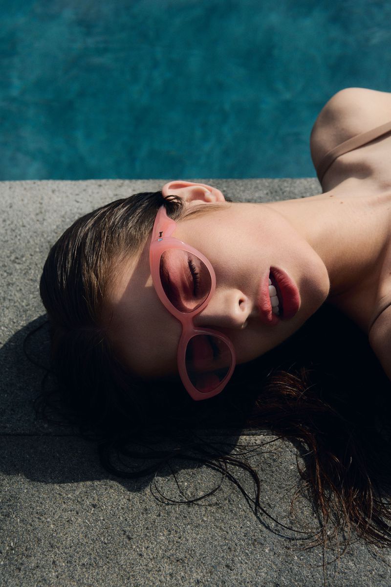 Close-up of a woman with dark hair wearing pink sunglasses, lying next to a blue swimming pool on a sunny day.