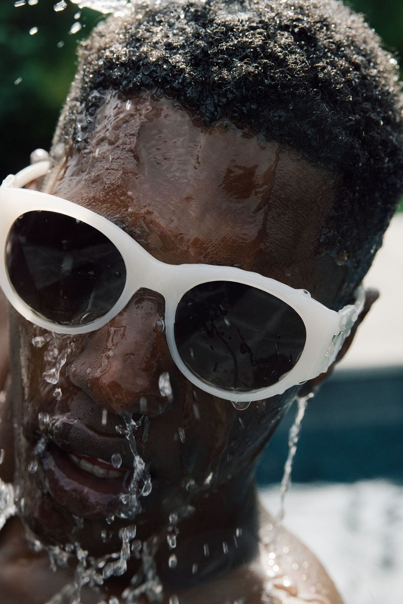 Close-up of an adult man with dark skin and short hair wearing white sunglasses while water is poured on his face, outdoor setting.