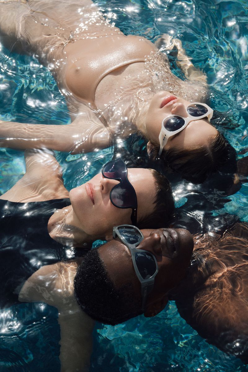 Three women of diverse ethnicities relax together floating in a shimmering blue swimming pool on a sunny day, wearing sunglasses.