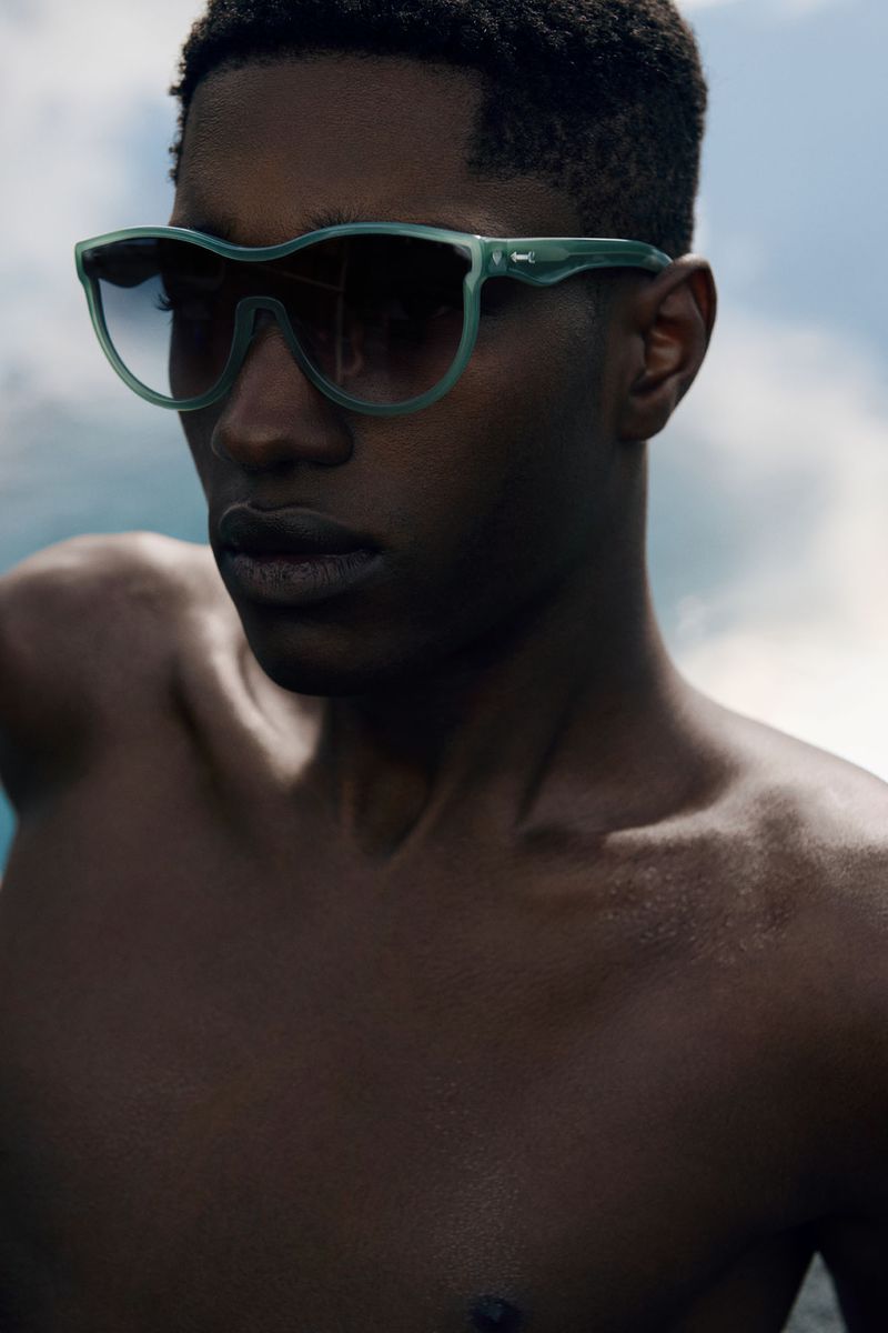 Close-up of a Black male model with sunglasses posing outdoors with a cloudy sky background, showcasing summer fashion and eyewear.