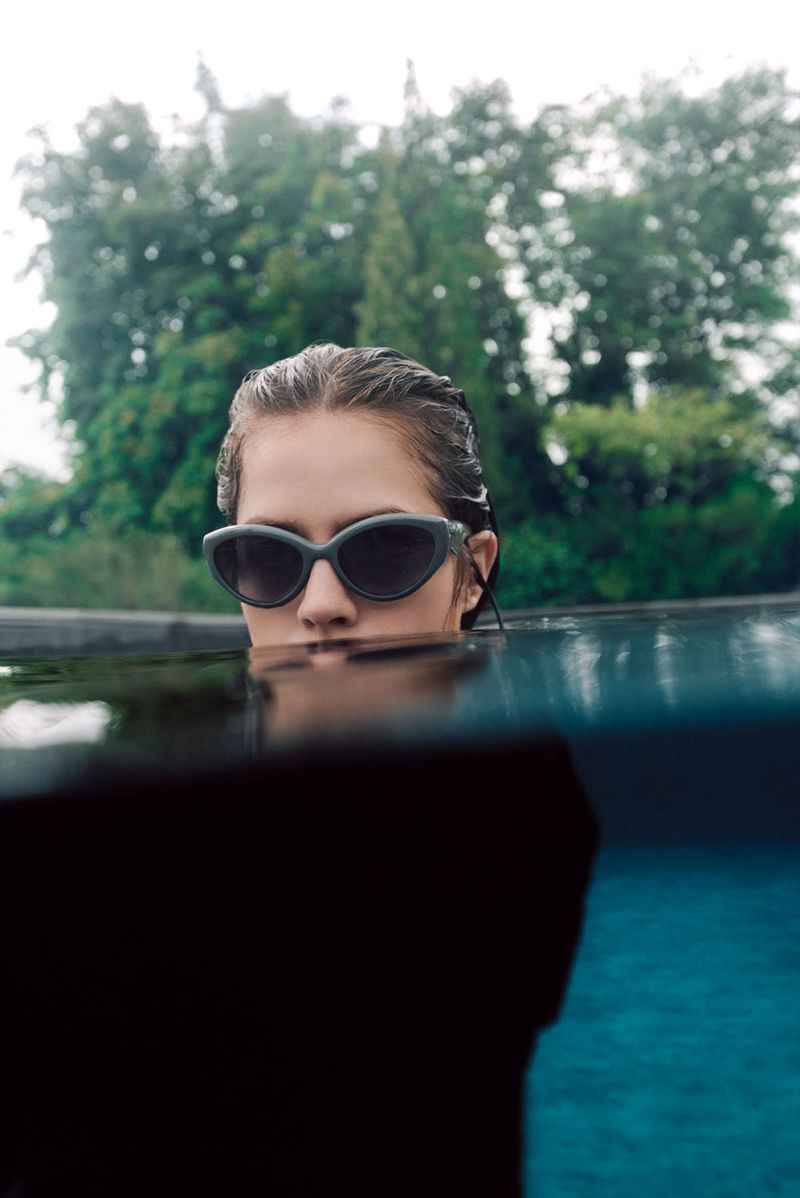 Portrait of a woman in sunglasses, chest-deep in water, against a backdrop of trees and sky, focusing on fashion and nature.