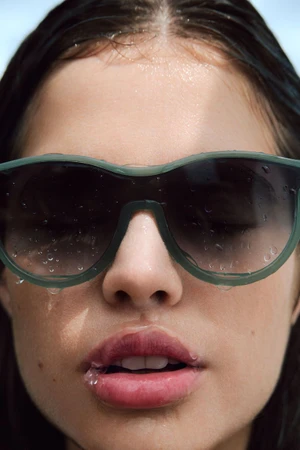 Close-up portrait of a model wearing dark green sunglasses covered in water droplets against a clear sky
