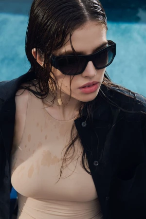 Model with wet hair wearing dark sunglasses and a beige top under an unbuttoned black shirt near a pool