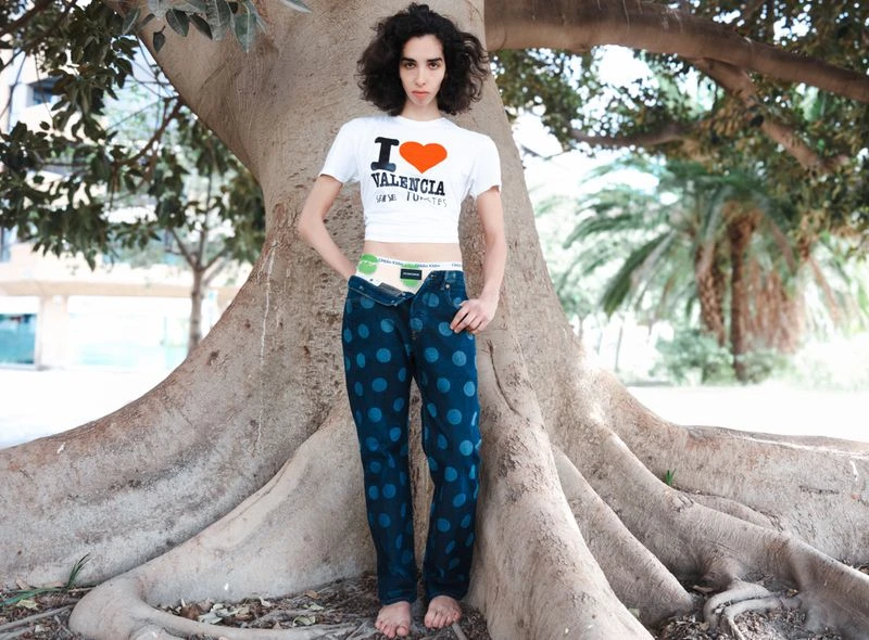 Young woman stands near a large tree with exposed roots wearing a white T-shirt and polka dot jeans, looking directly at the camera.