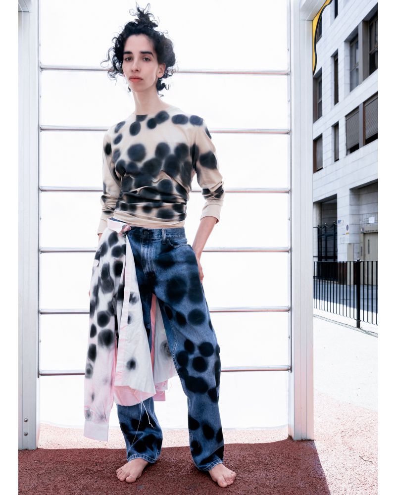 A barefoot woman stands in front of an outdoor structure wearing a polka dot shirt, dyed jeans, and a polka dot shirt tied around her waist.